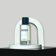 A bottle of Oudeis cologne displayed within a white, curved structure on a grey background.