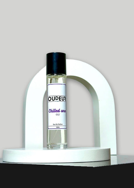 A bottle of unisex fragrance named 'OUDEUR' displayed on a white stand against a neutral background.